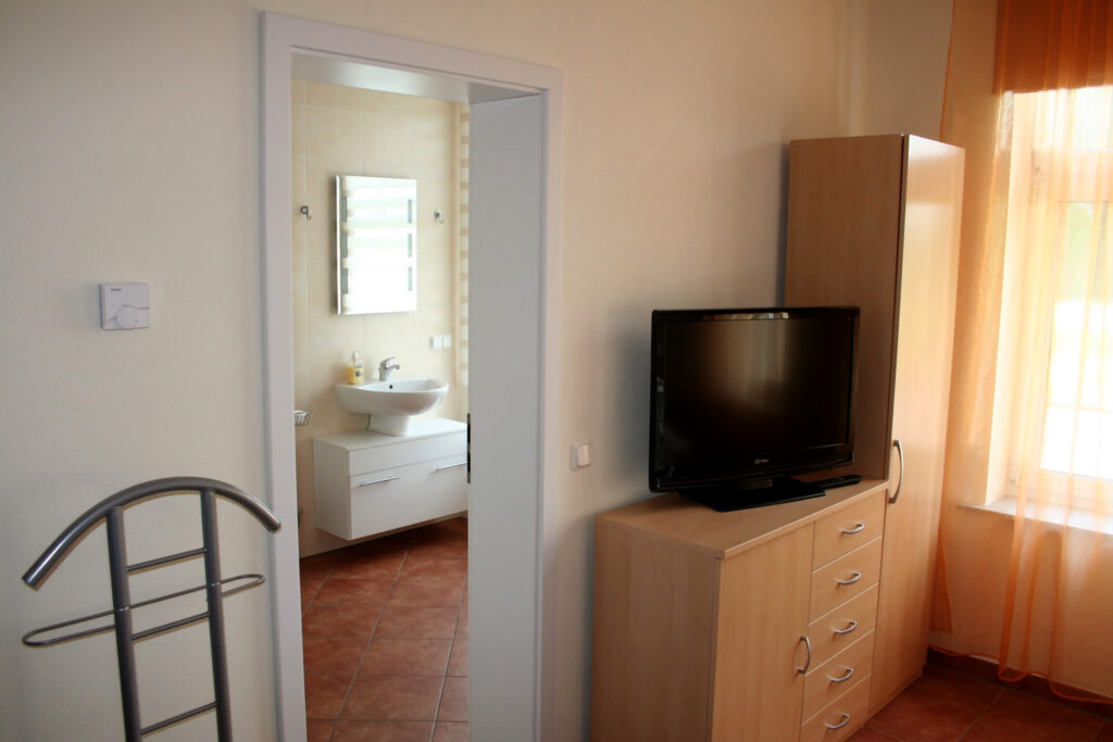 Schrank und Kommode mit Fernseher im Wohn- und Schlafbereich und Blick durch die offene Tür in das Badezimmer mit Waschbecken und Spiegel.
