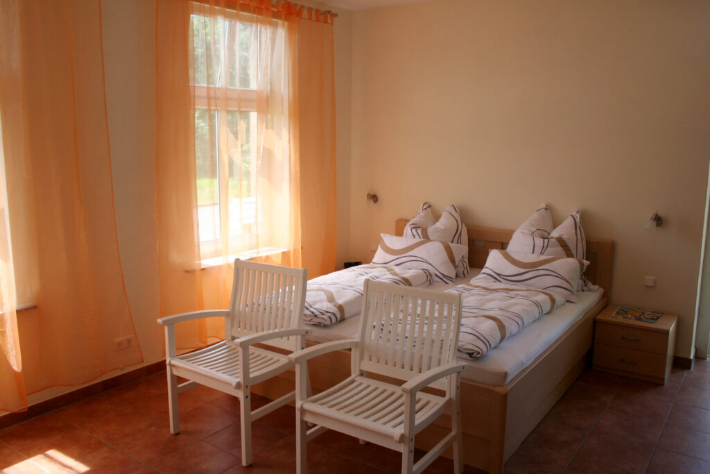 Blick auf den Schlafbereich der Ferienwohnung mit Doppelbett, gemusterter Bettwäsche, zwei weißen Stühlen am Fußende des Bettes, Nachttisch, Fenster mit Vorhängen und Tageslicht im Raum.