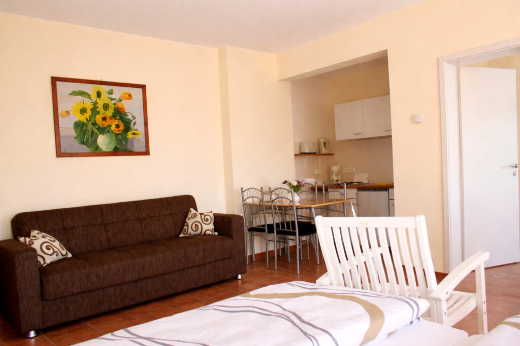 Blick über den Schlafbereich zum braunen Sofa mit Kissen, einem Wandbild mit Sonnenblumen, Esstisch mit Stühlen und offener Küchenzeile im Hintergrund.